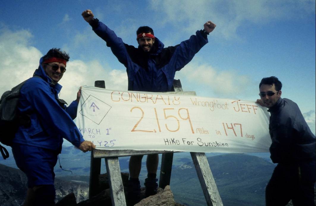 Three men holding a sign on top of a mountain.